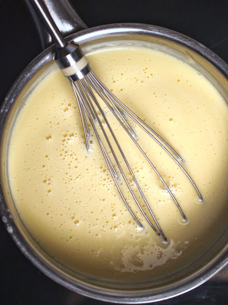 Thick, pale yellow ice cream custard rests in a small stainless steel pot with a wire whisk.