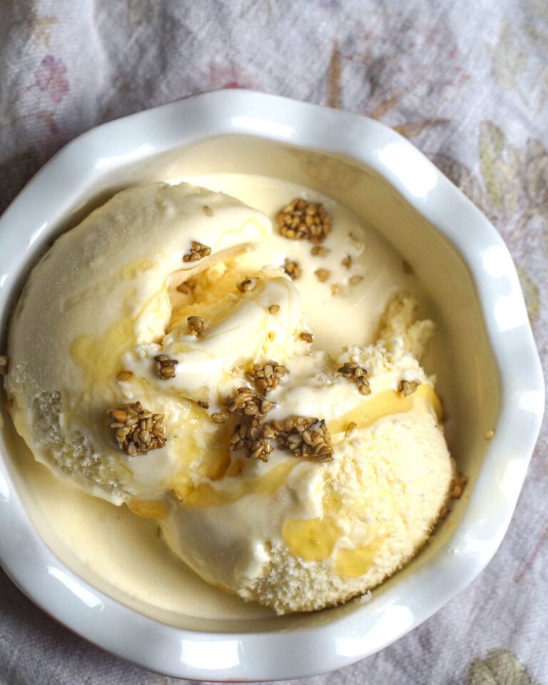 Salted Honey Ice Cream served with a drizzle of honey and a sprinkling of candied sesame crunch in a small white bowl with a fluted rim.