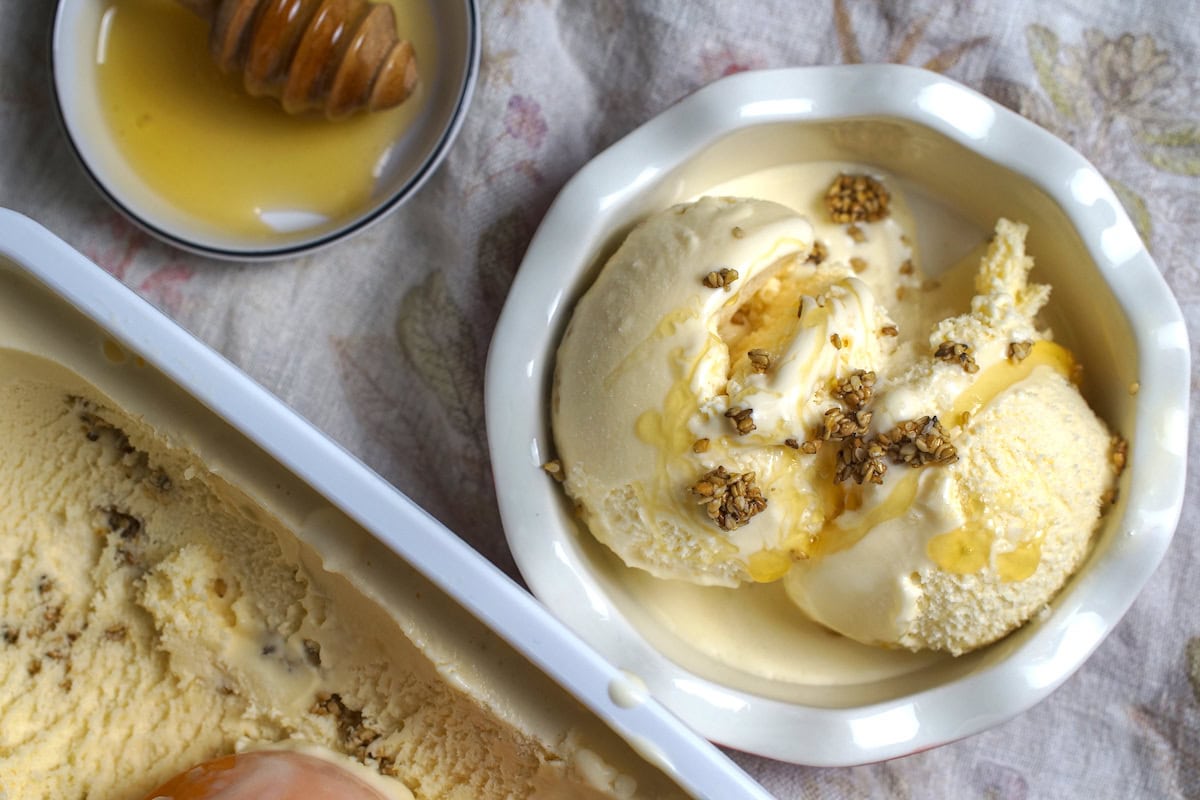 Two scoops of Salted Honey Ice Cream served with a drizzle of honey and a sprinkling of candied sesame crunch in a small white bowl with a fluted rim. The ice cream container and a small plate holding a honey spoon sit beside the bowl on a floral linen tea towel.