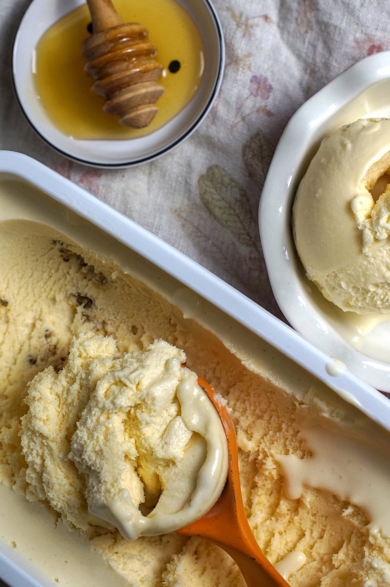 Salted Honey Ice Cream being scooped from a freezer container into a small white bowl with a fluted rim, next to a small plate holding a honey spoon. The filled ice cream scoop rests on top of the ice cream, showing the melting edges of a super creamy scoop.