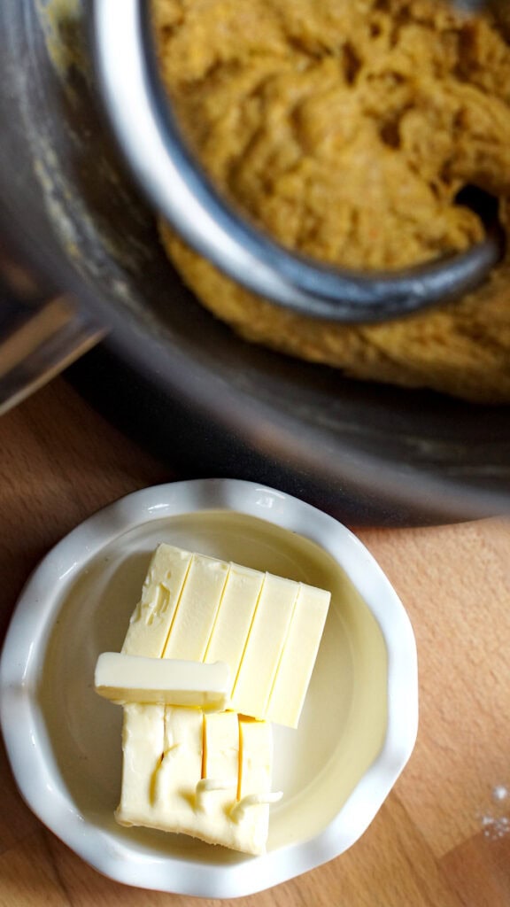 A small white bowl with fluted edge, holding cubed pieces of fridge-cold butter, sits next to a stand mixer, ready to be kneaded into the orange pumpkin sourdough brioche dough.