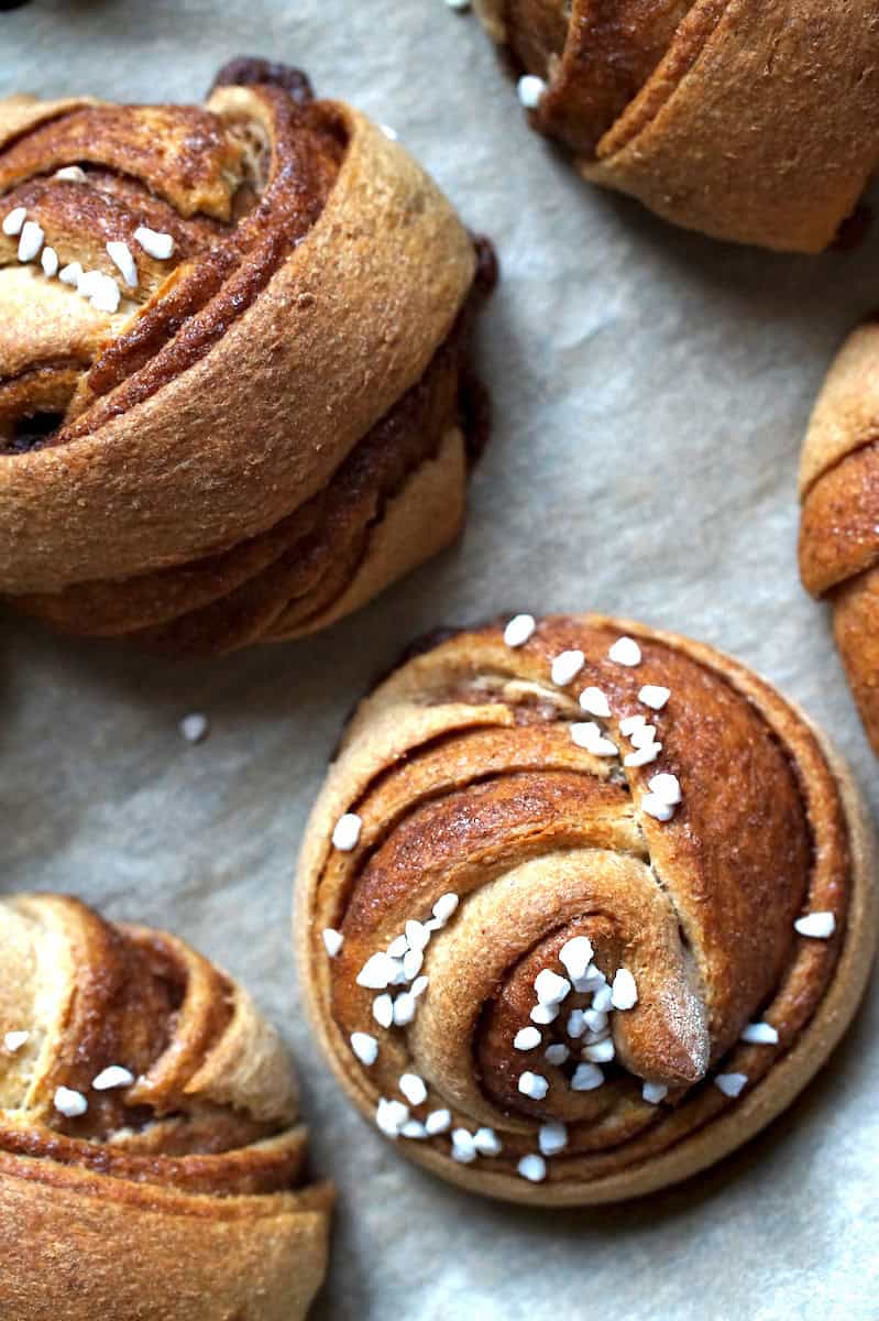 Closeup of a large, swirled, fluffy whole wheat Swedish cinnamon roll, topped with pearl sugar, sitting on a lined baking sheet.
