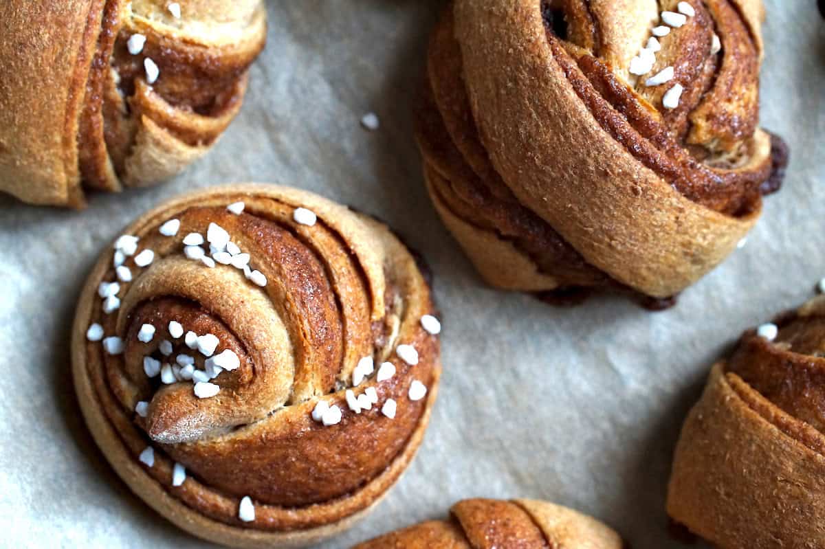 Closeup of a large, swirled, fluffy whole wheat Swedish cinnamon roll, topped with pearl sugar, sitting on a lined baking sheet.