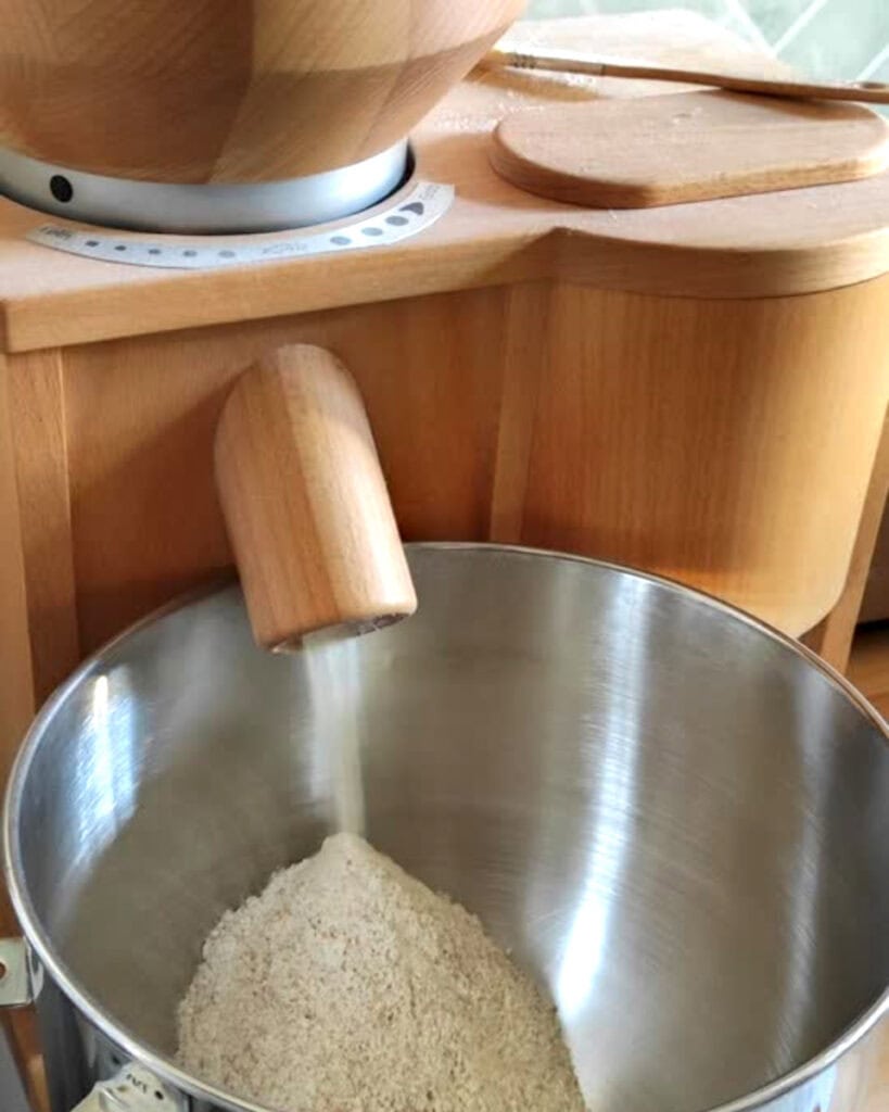 A stainless steel stand mixer bowl sitting underneath the chute of a wooden home grain bowl, catching fresh milled flour.