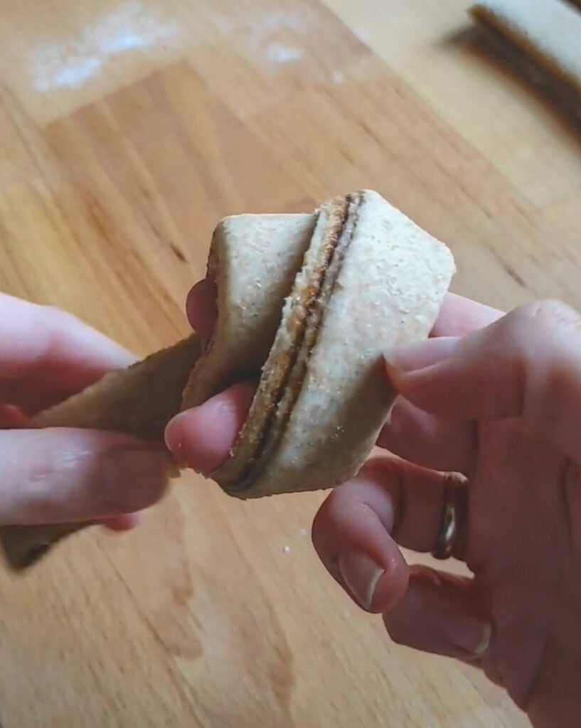 A strip of filled whole wheat cinnamon roll dough being wrapped around two fingers to shape a knotted bun.