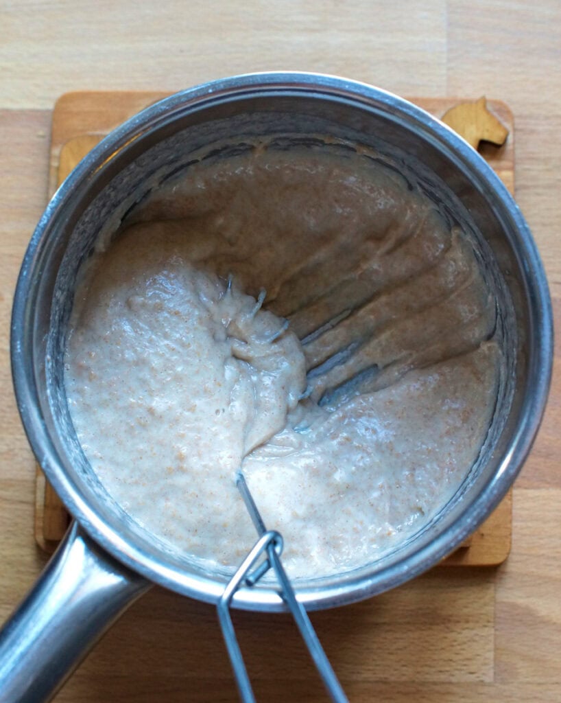 Thick cooked tangzhong roux holding the trail of a whisk in a small stainless steel pot on a wooden coaster.