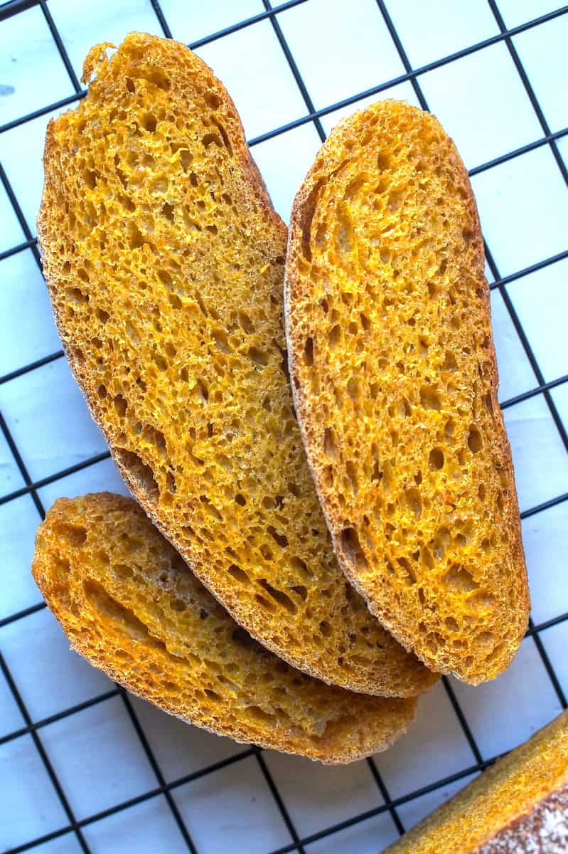 Three feathered out slices of sourdough pumpkin bread sitting on a black cooling rack on a chipped white wood surface.
