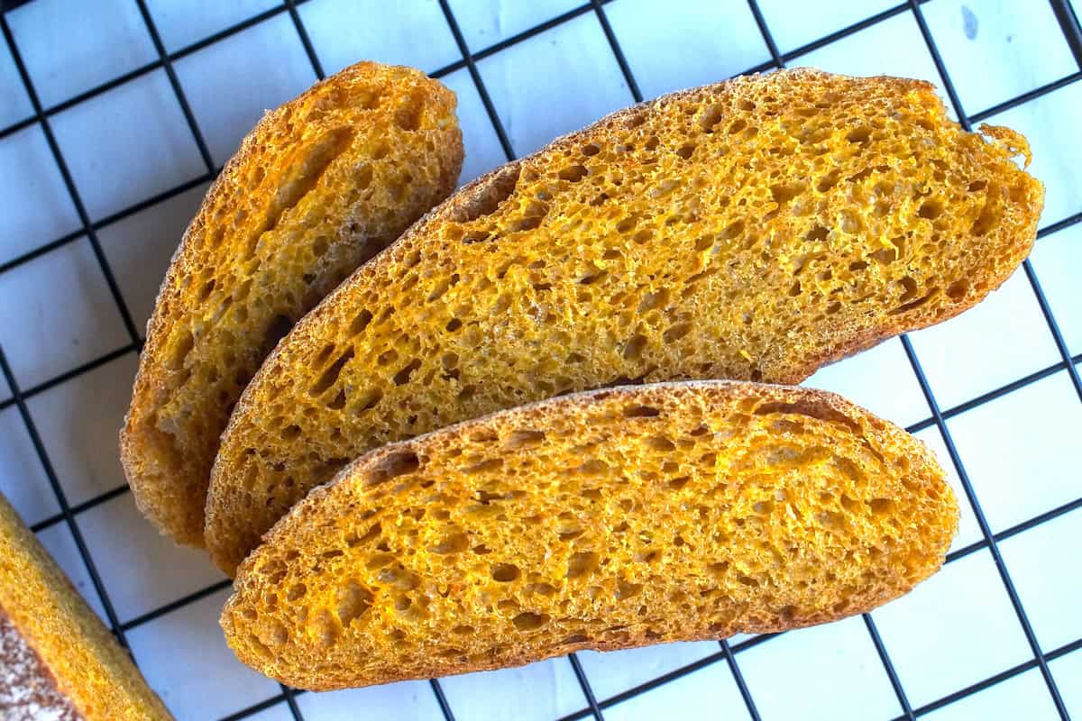 Three feathered out slices of sourdough pumpkin bread sitting on a black cooling rack on a chipped white wood surface.