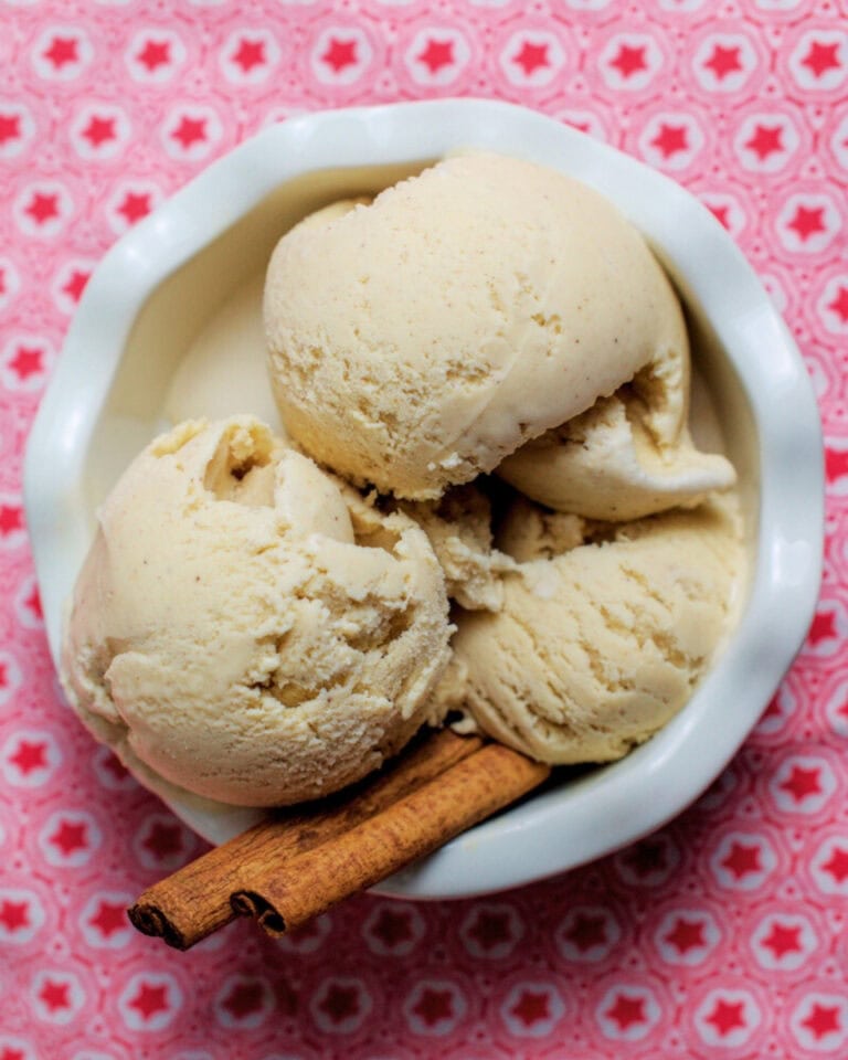 Three scoops of creamy cinnamon ice cream in a white fluted bowl, garnished with cinnamon sticks, on a red-star tea towel.
