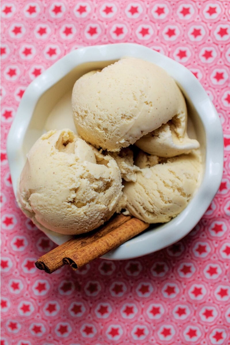Three scoops of creamy cinnamon ice cream in a white fluted bowl, garnished with cinnamon sticks, on a red-star tea towel.
