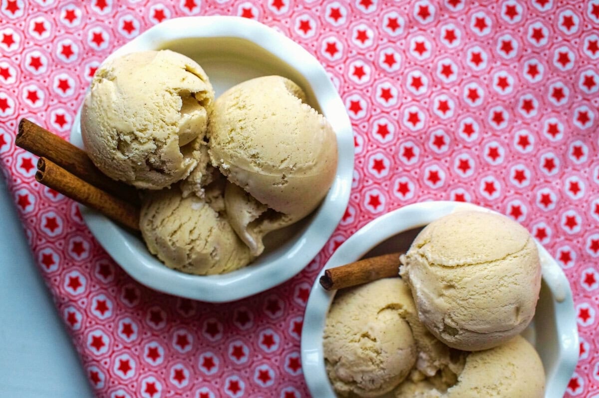 Two bowls with three scoops each of creamy cinnamon ice cream in a white fluted bowl, garnished with cinnamon sticks, on a red-star tea towel.