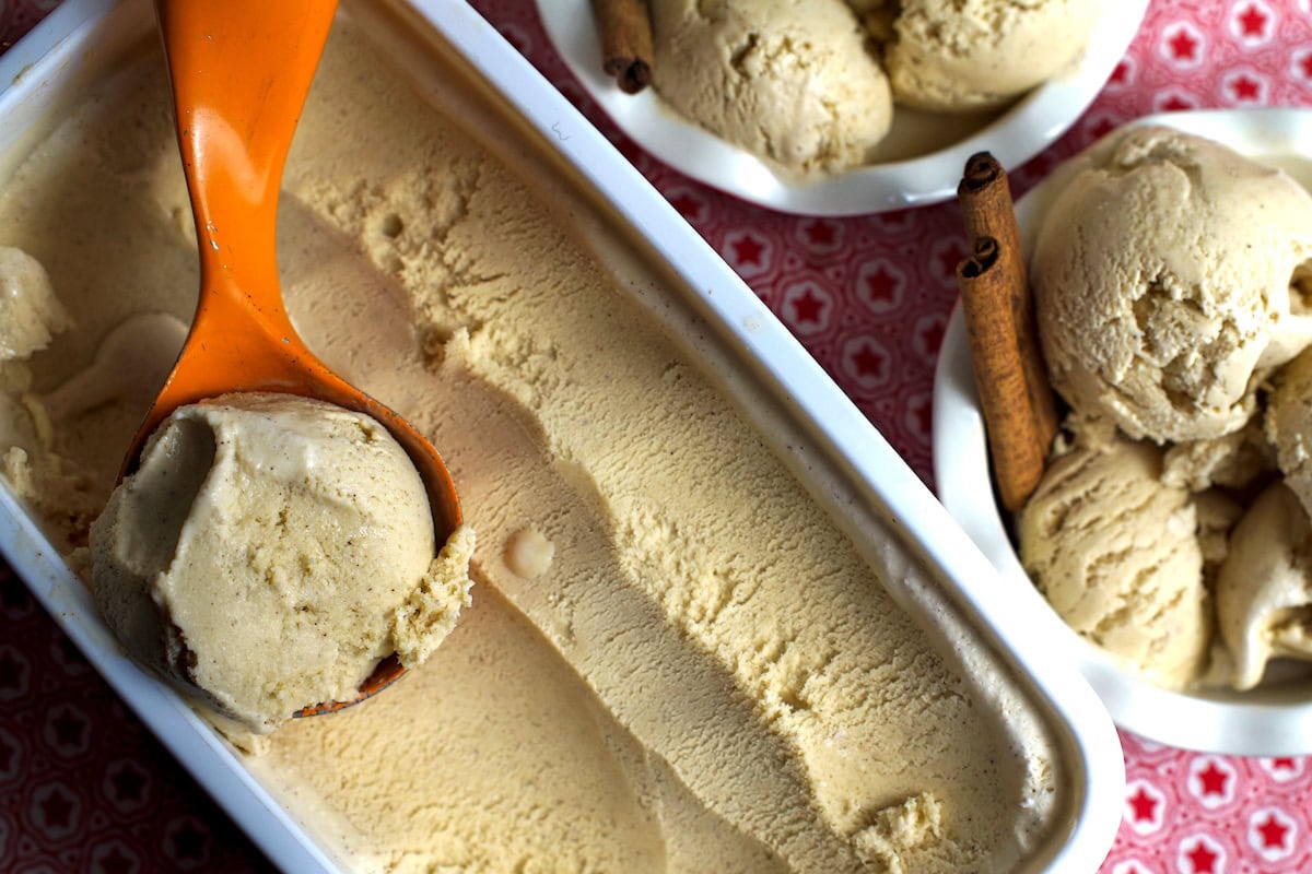 Two white fluted bowls, each with three scoops of cinnamon ice cream, alongside a container holding a scoop and another scoop on a spoon, placed on a red-star tea towel.