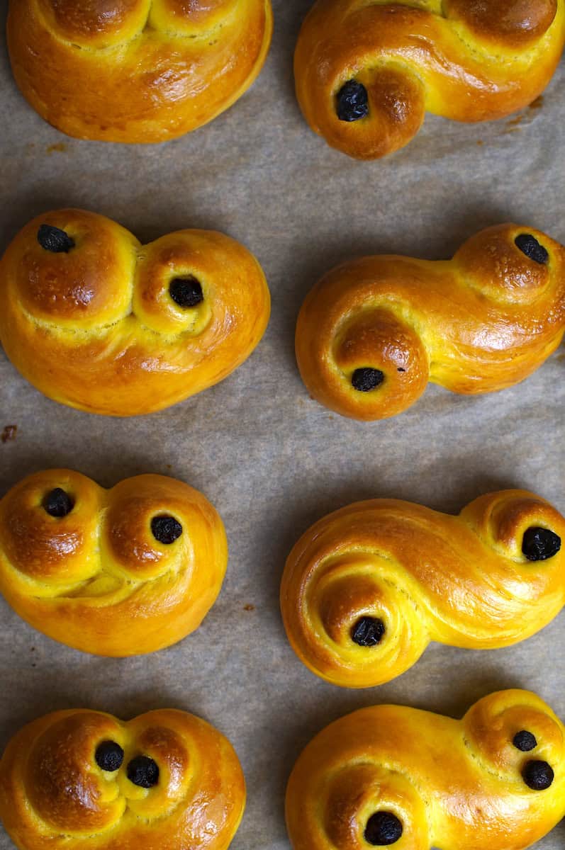 Traditionally shaped sourdough lussekatter sitting on a lined baking sheet, decorated with raisins and baked to golden perfection.