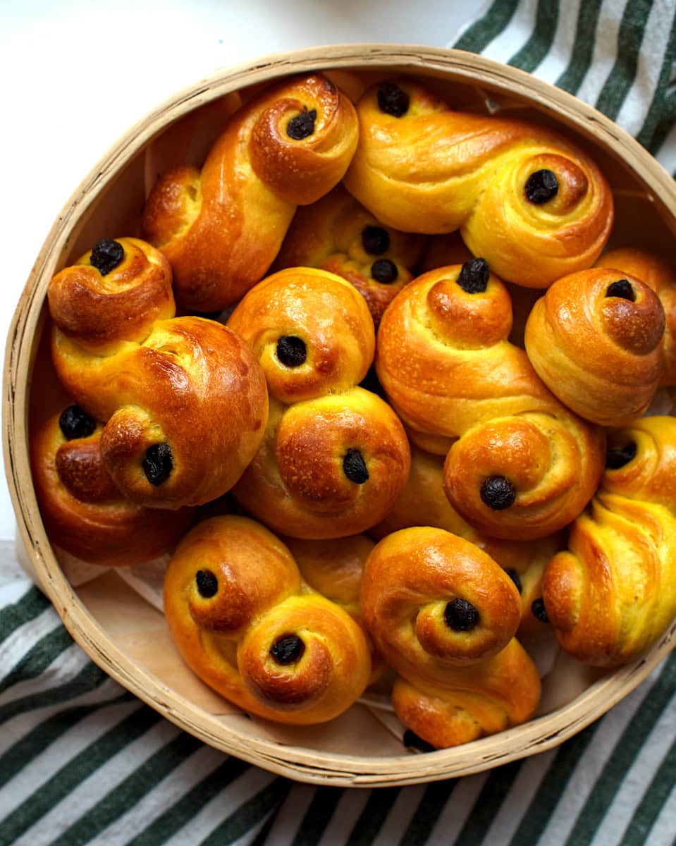 A basket filled with freshly baked sourdough lussekatter, or Swedish saffron buns, is presented on a green and white striped tea towel on a chipped white wooden surface.