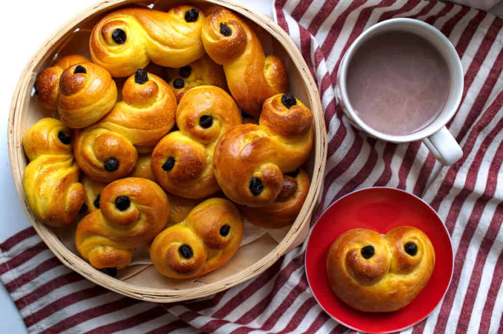 A basket filled with freshly baked sourdough lussekatter, or Swedish saffron buns, is presented alongside a red plate featuring one of the saffransbröd, with a cup of hot cocoa nearby, all set against a red and white striped surface.