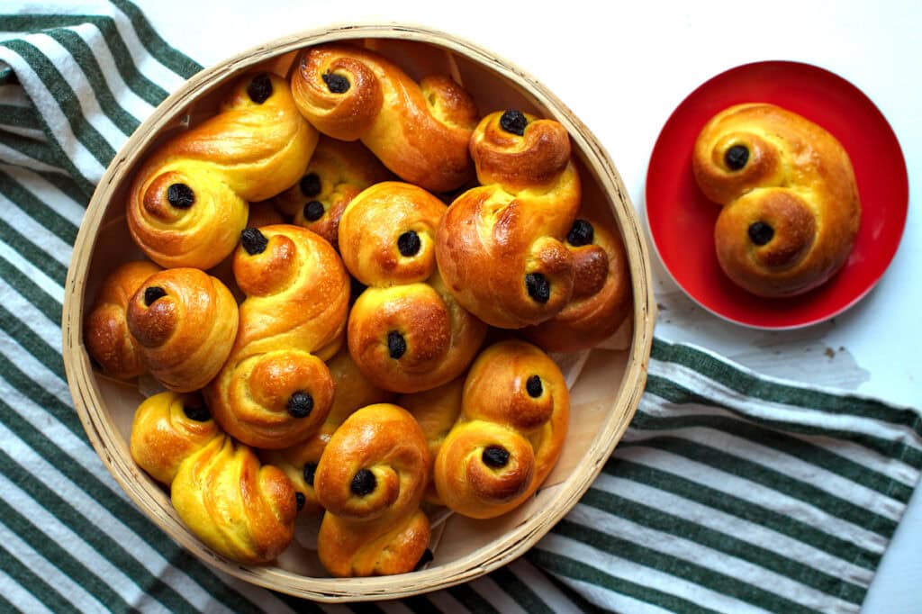 A basket filled with freshly baked sourdough lussekatter, or Swedish saffron buns, is presented alongside a red plate featuring one of the saffransbröd, all set against a green and white striped surface.