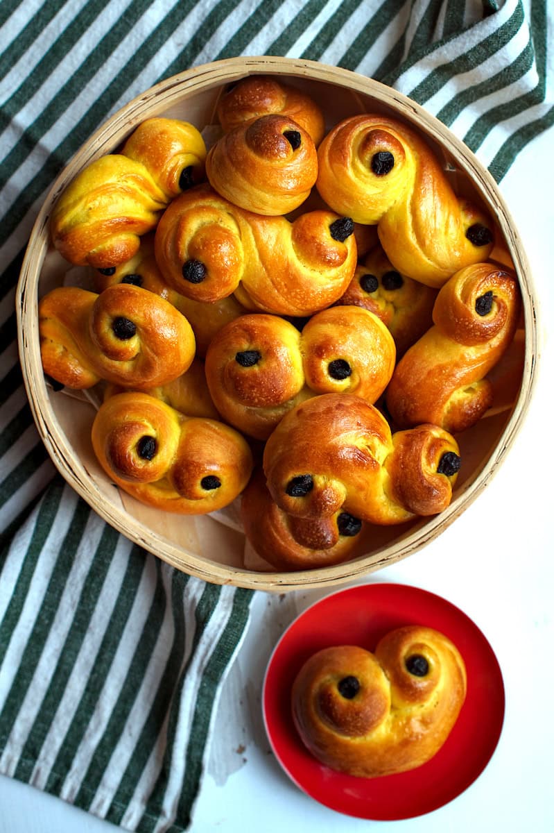 A basket filled with freshly baked sourdough lussekatter, or Swedish saffron buns, is presented alongside a red plate featuring one of the saffransbröd, all set against a green and white striped surface.