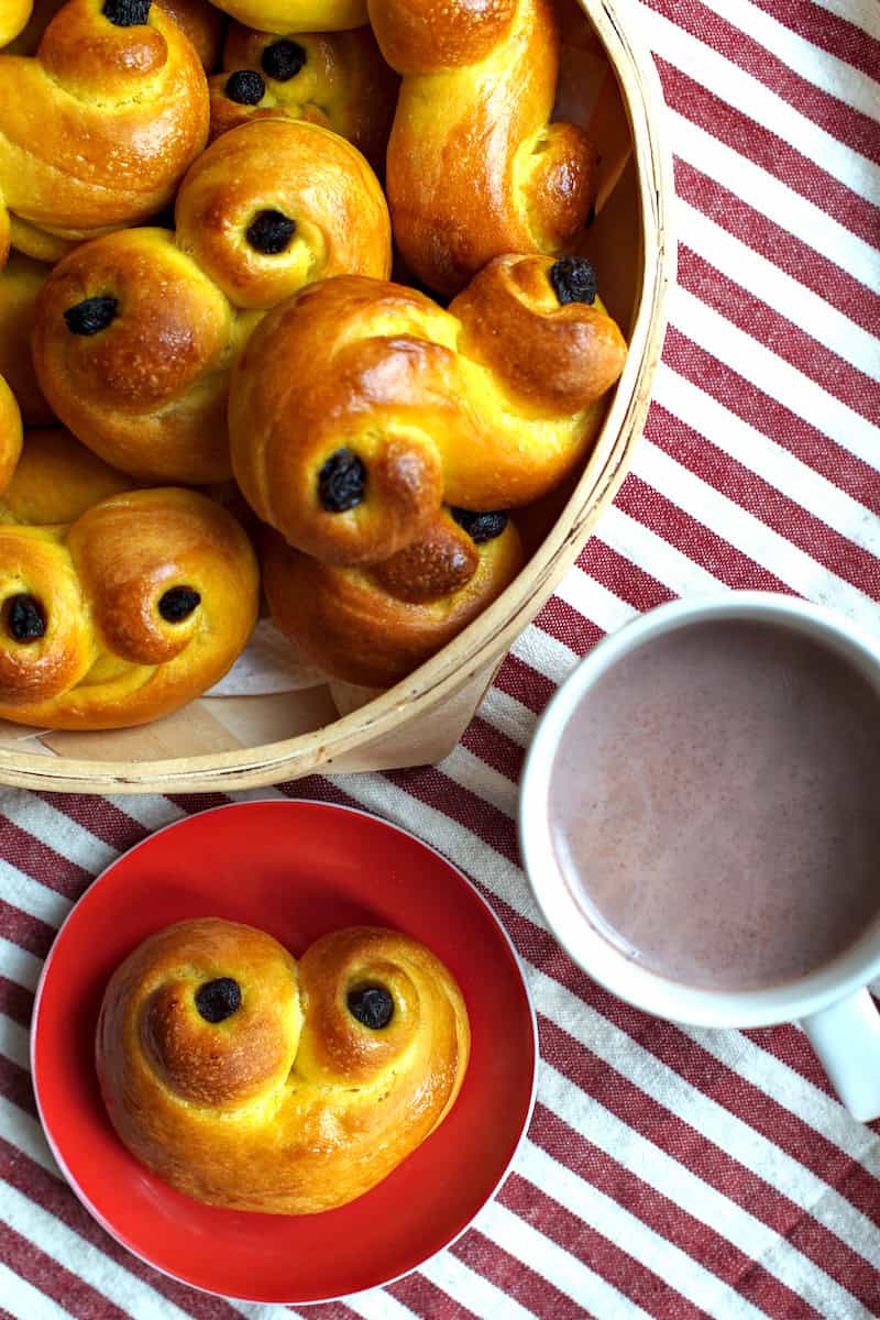 A basket filled with freshly baked sourdough lussekatter, or Swedish saffron buns, is presented alongside a red plate featuring one of the saffransbröd, with a cup of hot cocoa nearby, all set against a red and white striped surface.