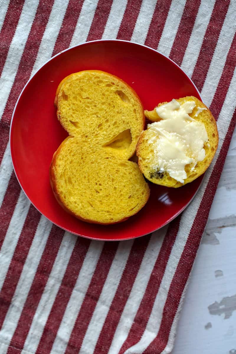A split sourdough saffron bun on a small red plate, sitting on a striped tea towel on a chipped white wooden surface, exposing the even, orange colored crumb. One piece of the lussekatter is spread with butter.