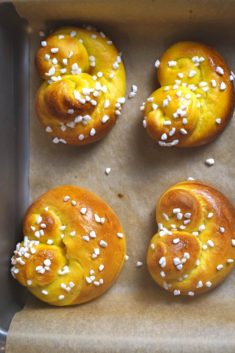 Four baked sourdough lussekatter on a small baking sheet lined with baking paper, topped with bright white pearl sugar as an alternative to traditional raisins.