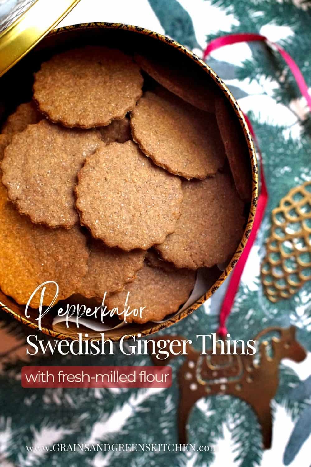 A festive, golden cookie tin is filled to the top with dark, crispy freshly milled pepparkakor. The tin is set out on a table runner printed with dark, evergreen leaves and branches, next to a golden Scandinavian Christmas horse decoration.