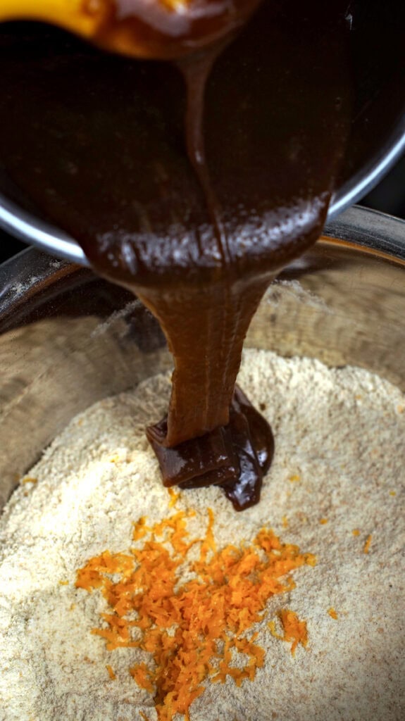 Dark, glossy syrup-butter-mix is poured nto a bowl filled with fresh milled soft wheat flour and grated orange zest.