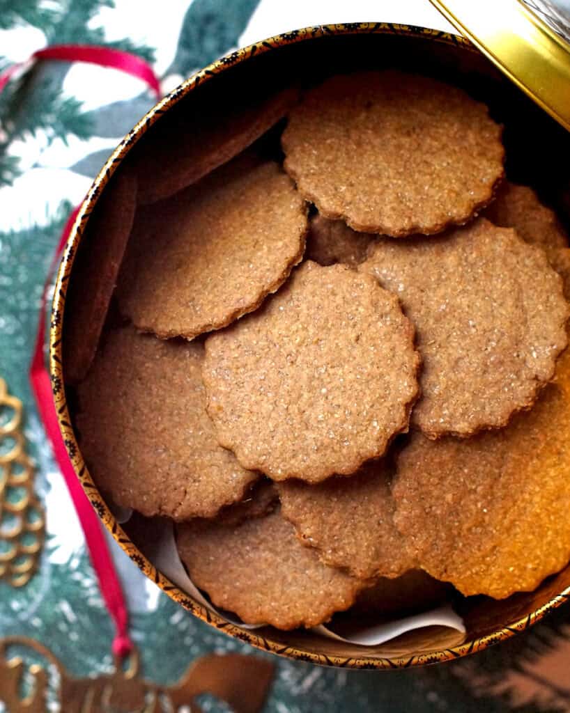 A festive, golden cookie tin is filled to the top with dark, crispy freshly milled pepparkakor. The tin is set out on a table runner printed with dark, evergreen leaves and branches, next to a golden Scandinavian Christmas horse decoration.
