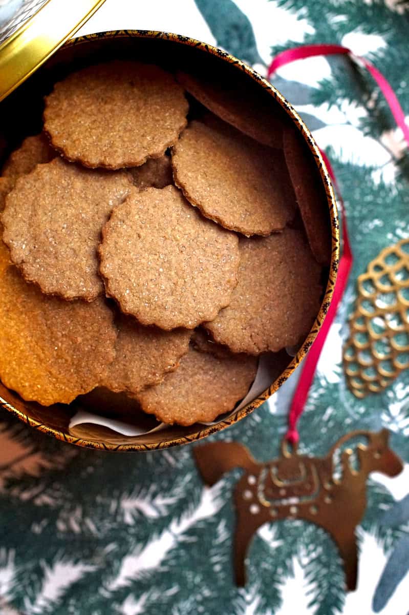 A festive, golden cookie tin is filled to the top with dark, crispy freshly milled pepparkakor. The tin is set out on a table runner printed with dark, evergreen leaves and branches, next to a golden Scandinavian Christmas horse decoration.
