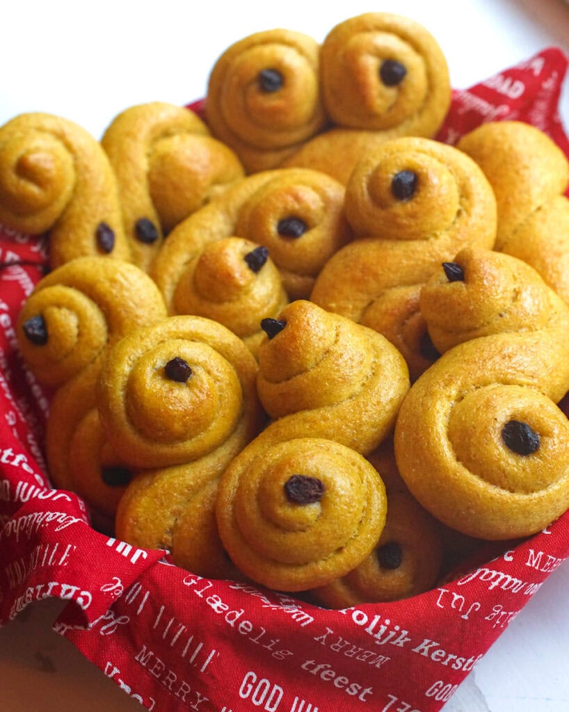 A basket lined with a red Christmas-themed cloth napkin is filled with tender, golden whole wheat sourdough saffron buns, made with freshly milled flour. The buns are twisted into different shapes and decorated with raisins.
