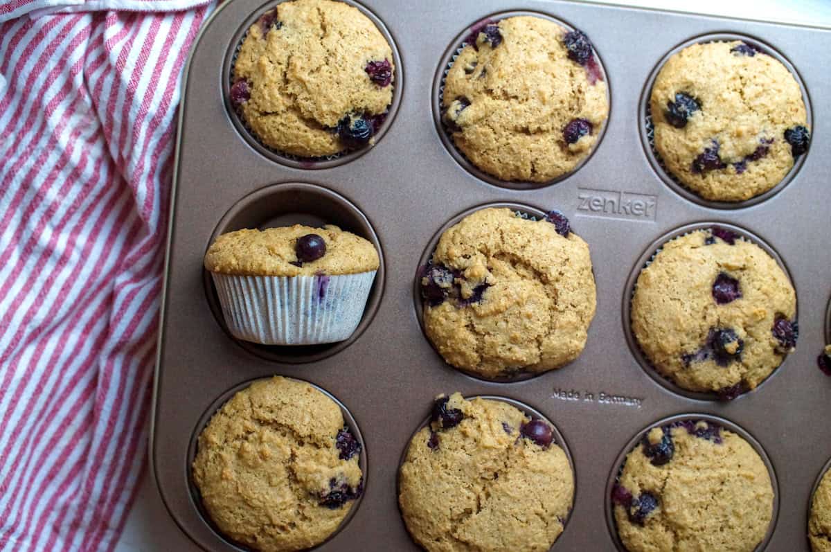 Top-down view of baked sourdough blueberry muffins in white liners, one muffin flipped on its side to showcase its perfectly domed top, next to a striped tea towel.
