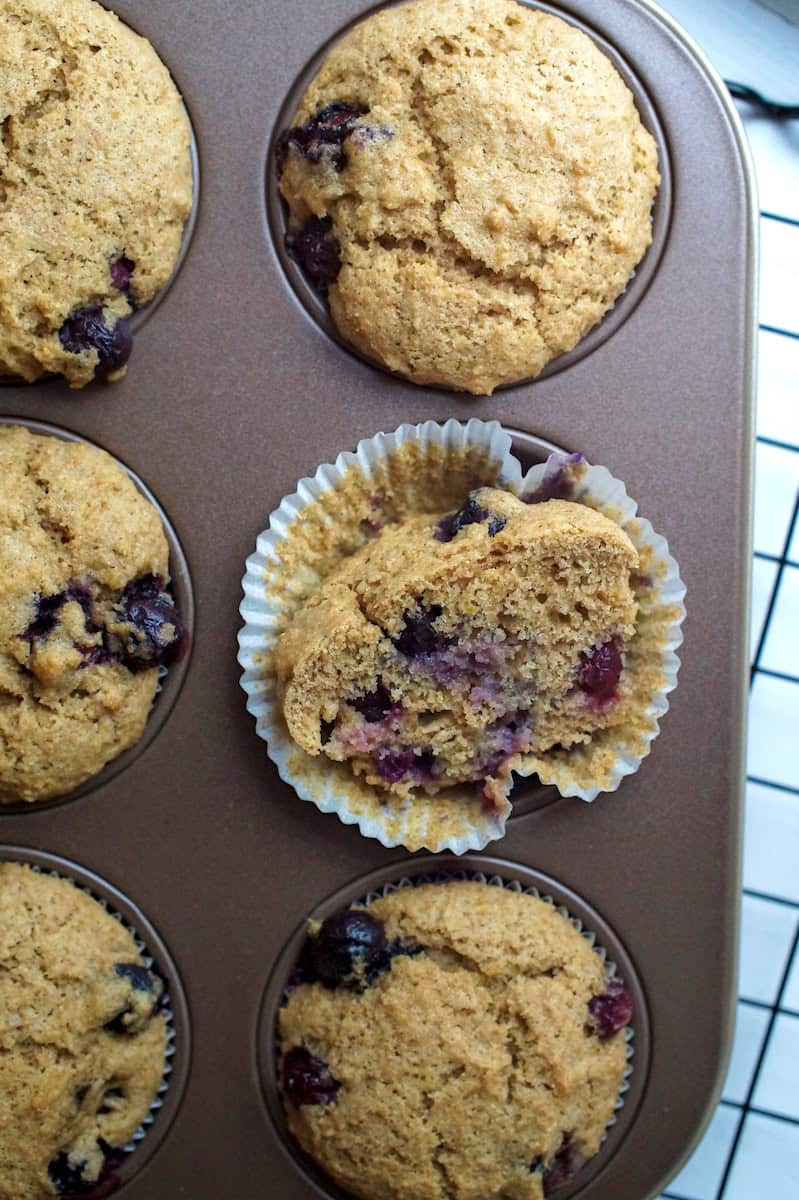 Top-down view of sourdough blueberry muffins in white liners, with one muffin split open to reveal its tender, fluffy interior, resting on the unwrapped liner in the muffin tin.