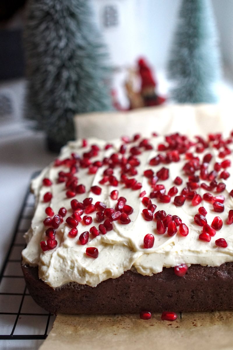 Orange sourdough gingerbread on a cooling rack, topped with white frosting and red pomegranate seeds, with festive Christmas decorations in the background.
