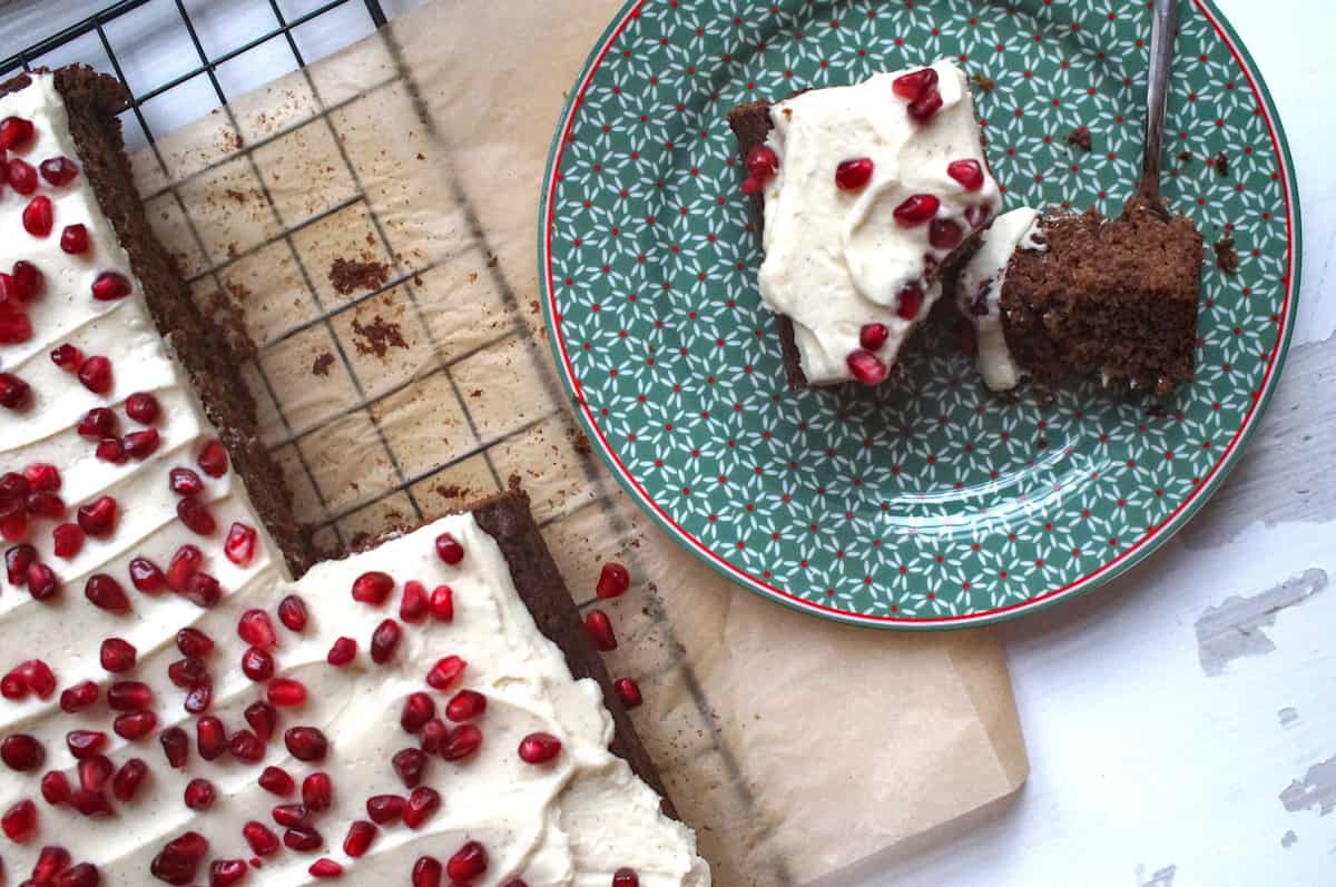 A slice of orange sourdough gingerbread sits on a dark green festive plate, topped with creamy vanilla cream cheese frosting and jewel-like pomegranate seeds. The plate rests on a weathered white wooden board, next to the black cooling rack holding the full cake on brown parchment paper.