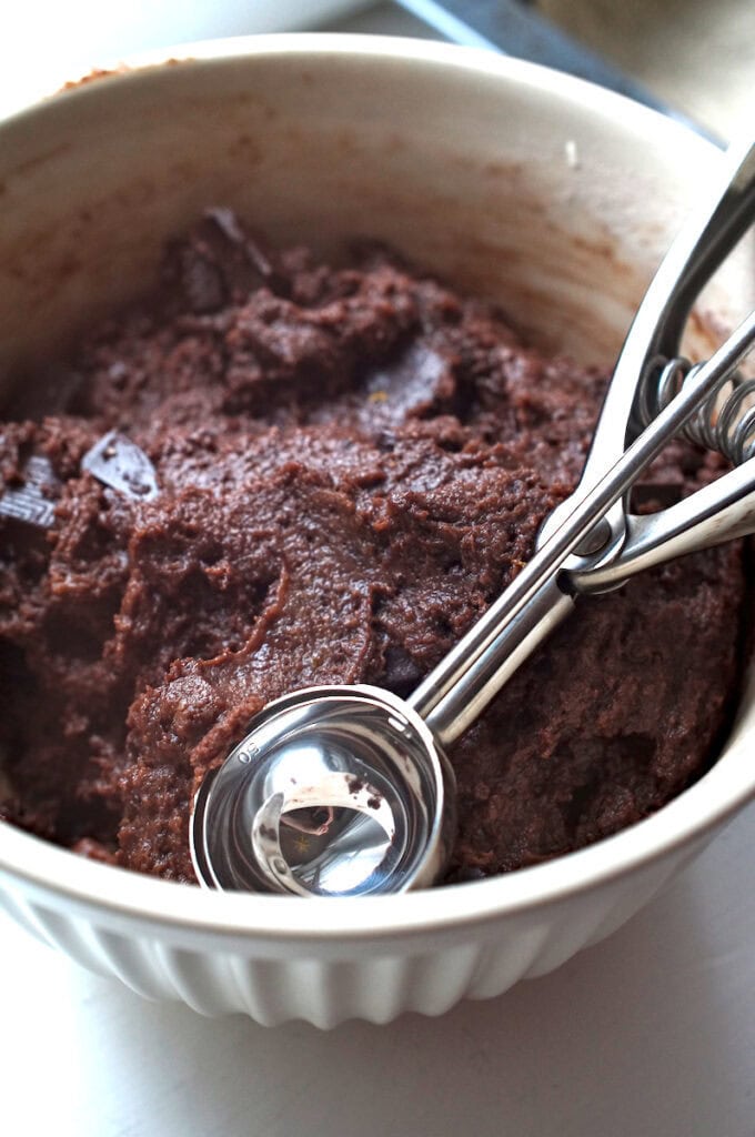 Close-up of dark, glossy double chocolate sourdough cookie batter in a cream ceramic bowl, with large chocolate chunks glistening in the light and a silver cookie scoop resting inside, marked size 50.