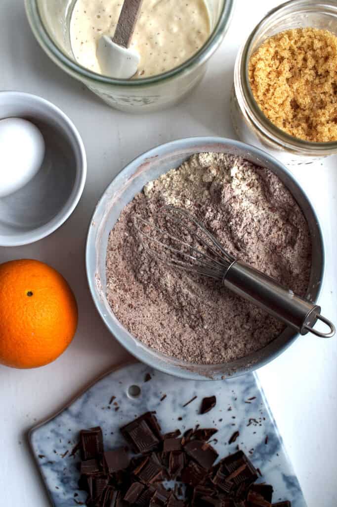 Top-down view of double chocolate sourdough cookie ingredients: blue bowl of dry mix with whisk, brown sugar, sourdough discard, egg, orange, and chopped chocolate.