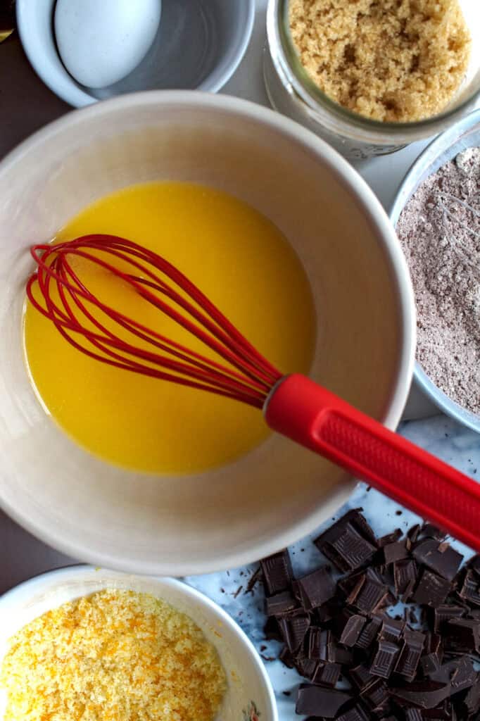 Cream bowl of melted butter with red whisk, surrounded by chopped chocolate, orange sugar, brown sugar, dry ingredients, and a whole egg.