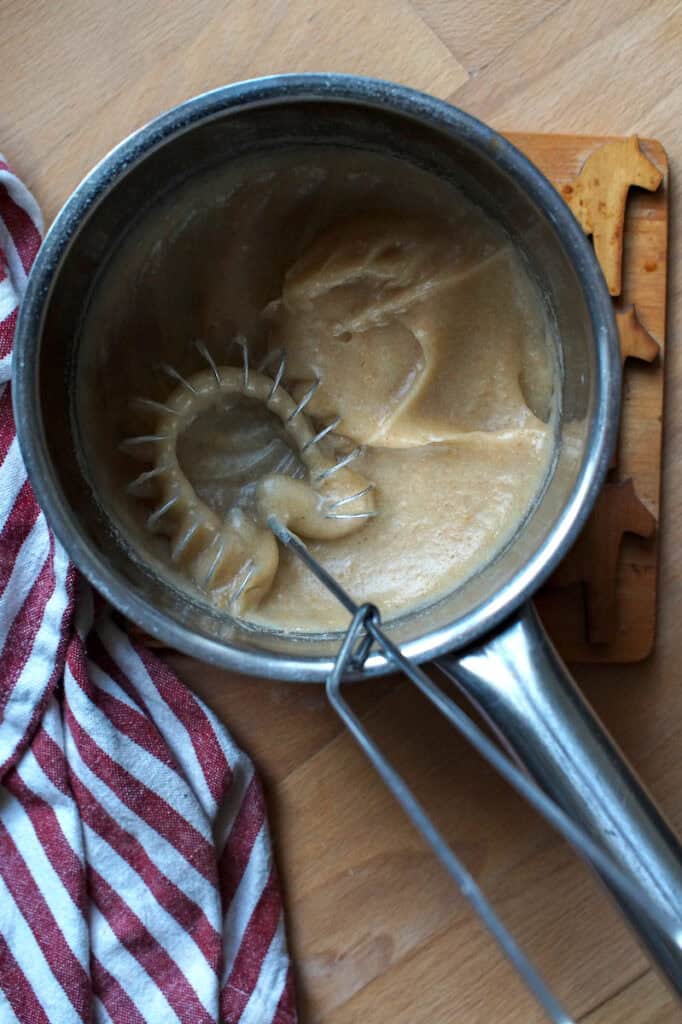 Thick tangzhong roux made with fresh milled whole wheat flour in a small silver saucepan with flat whisk on a wooden coaster.