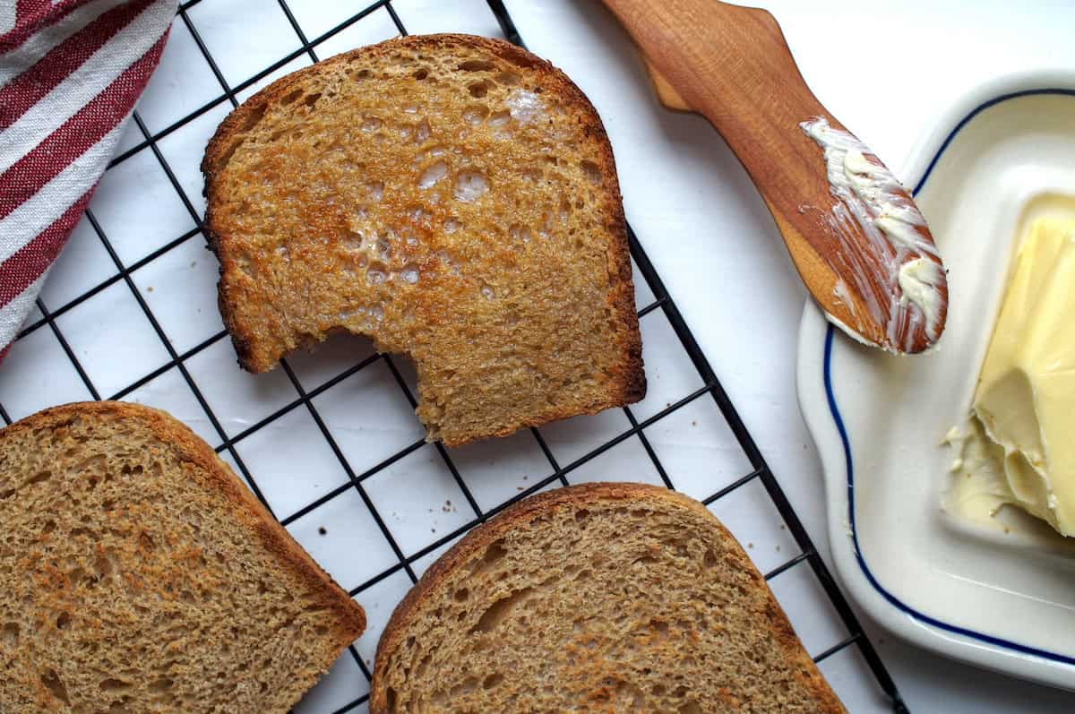 Three toasted slices of European-style whole wheat sourdough sandwich bread with fresh milled flour, one buttered with a bite.