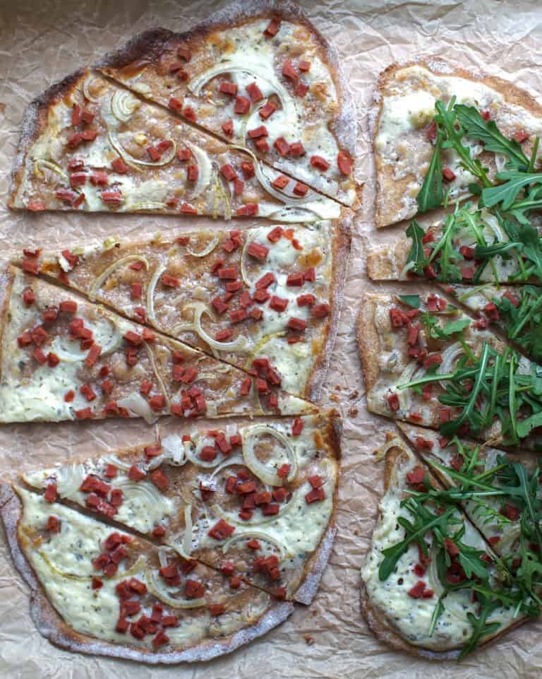 Top-down view of two crispy baked flammkuchen on crinkled parchment paper, one topped with dark green arugula.