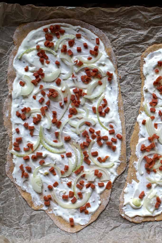 Overhead view of rolled-out sourdough flammkuchen dough on a parchment lined baking sheet, topped with sour cream, thinly sliced onions, and chopped bacon.