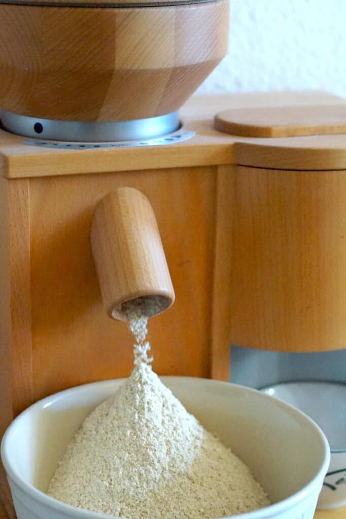 Freshly milled flour is falling from the spout of a wooden home grain mill into a ceramic bowl underneath.