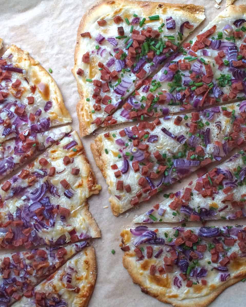 Overhead view of two crispy flammkuchen cut into wedges on parchment paper, one sprinkled with fresh green chives, showing thin, golden-brown Alsatian flatbread with sour cream, onions, and veggie bacon