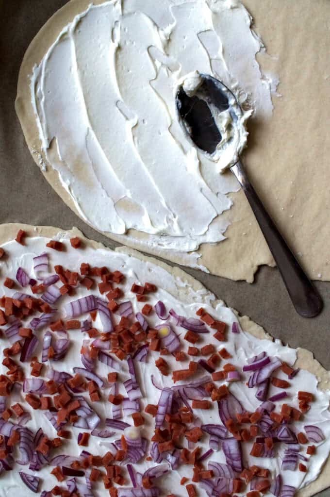 Two flammkuchen on a baking sheet before baking, one fully assembled with sour cream, red onions, and veggie bacon, the other being topped with sour cream with a spoon resting on the dough, ready to bake