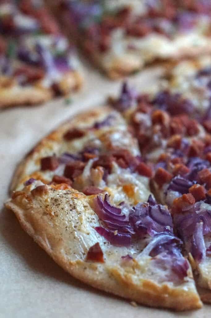 Close-up of a crispy, blistered edge of a flammkuchen topped with red onions, veggie bacon, and a thin layer of sour cream, highlighting the golden-brown, paper-thin crust