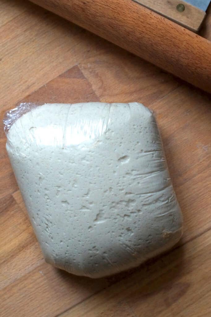 Puffy sourdough discard flammkuchen dough pressing against plastic wrap on a wooden counter, next to a rolling pin and bench scraper, showing a well-fermented, elastic texture