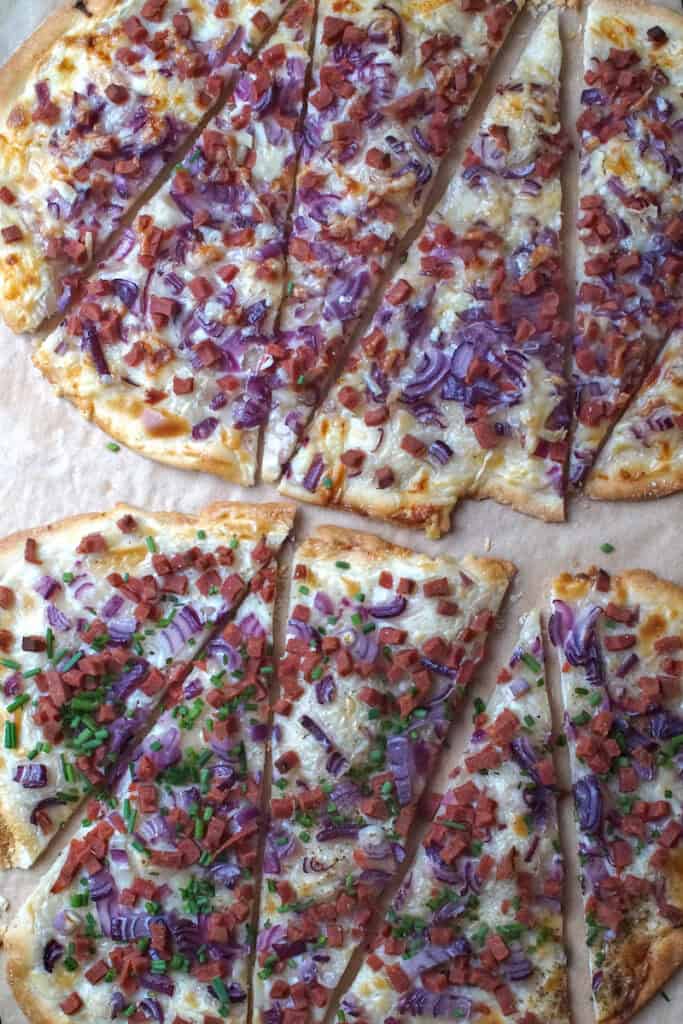 Angled overhead view of two crispy flammkuchen cut into wedges on parchment paper, one sprinkled with fresh green chives, showing thin, golden-brown Alsatian flatbread with sour cream, onions, and veggie bacon