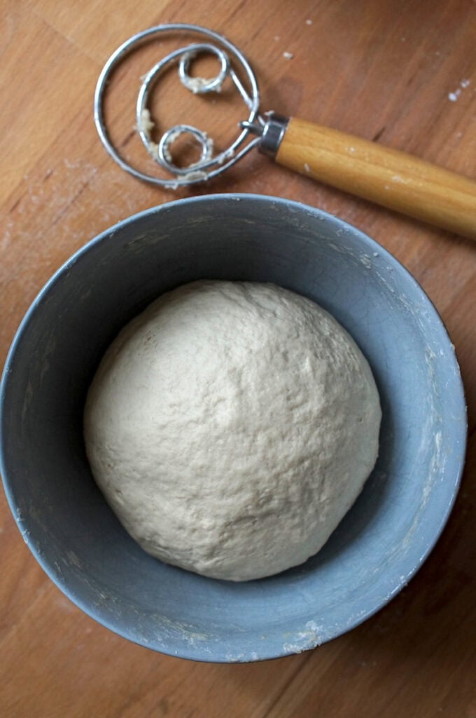 Top-down view of a rustic blue bowl containing smooth, elastic sourdough discard flammkuchen dough, ready to rest or roll, with a Danish dough whisk on a wooden counter