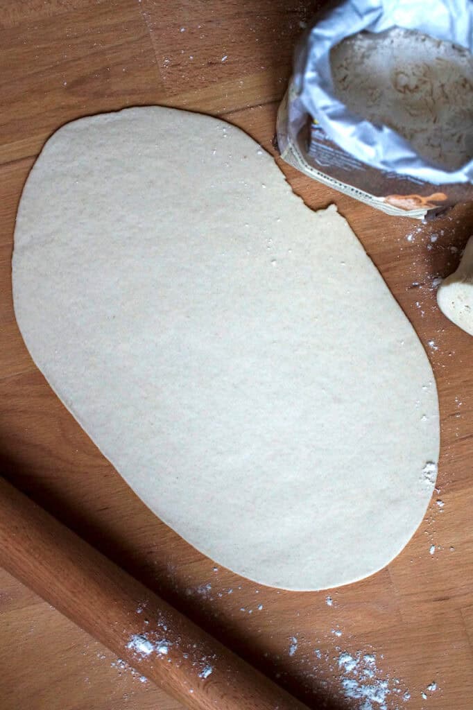 Top-down view of paper-thin flammkuchen dough rolled out on a wooden counter, with a rolling pin, a bag of flour, and a light dusting of flour, ready for baking a crispy Alsatian flatbread