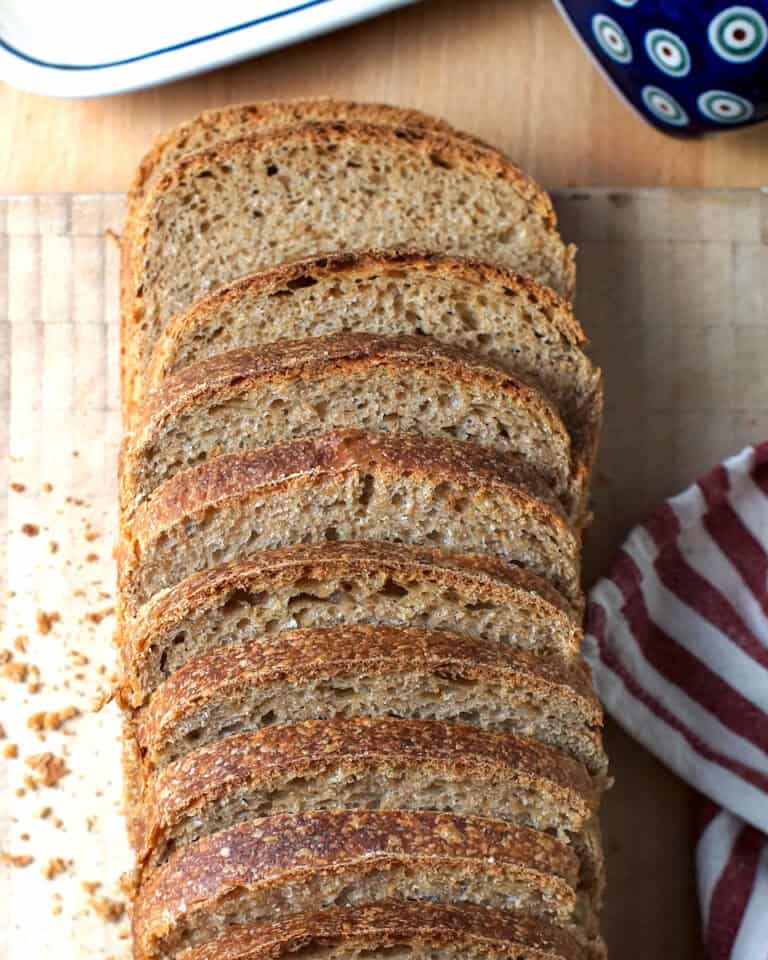 Top-down view of a whole brioche-style sourdough sandwich bread loaf made with fresh milled whole wheat flour, sliced and fanned on a cutting board, with tea towel and butter dish nearby.