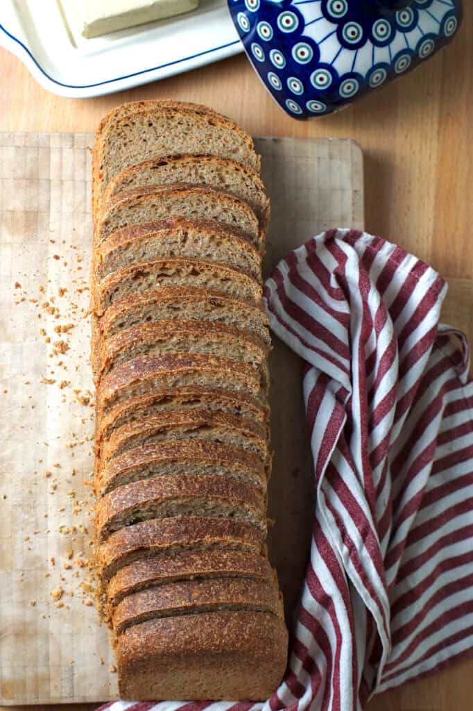 Top-down view of a whole brioche-style sourdough sandwich bread loaf made with fresh milled whole wheat flour, sliced and fanned on a cutting board, with tea towel and butter dish nearby.